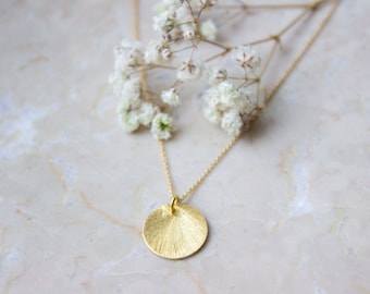 Könnte beinhalten: Eine goldene Halskette mit einem runden, strukturierten Anhänger. Der Anhänger hat ein Sonnenstrahlen-Design. Die Halskette liegt auf einer weißen Marmorfläche mit weißen Blumen.