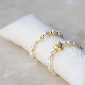 Könnte beinhalten: Zwei zarte Perlenarmbänder mit goldenen Akzenten. Ein Armband hat einen kleinen goldenen Herzanhänger.