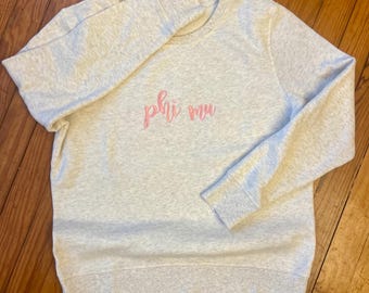 May include: A light gray sweatshirt with the embroidered text "phi mu" in pink script. The crew neck sweatshirt has long sleeves and a ribbed hem and cuffs. The garment is made of a soft, comfortable material.