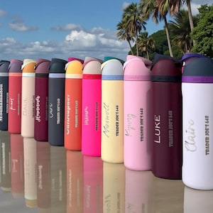May include: A row of Owala insulated water bottles in a range of colours, including pink, red, and white. Each bottle has a name printed on it, with the Trader Joe's 640 logo. The bottles are arranged on a reflective surface.