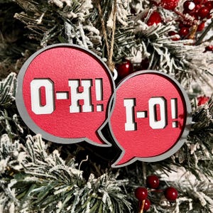 May include: Two red and gray speech bubble ornaments with the text "O-H!" and "I-O!" in white block letters. The ornaments are hanging on a Christmas tree with red berries.