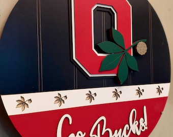 May include: A circular wooden wall sign featuring a large red block letter O with white and black borders. A green buckeye leaf and nut are attached to the O. Below, a white stripe with leaf cutouts sits above a red section with Go Bucks! text.