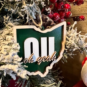 May include: A green and white wooden ornament shaped like the state of Ohio with the letters "OU" and the text "oh yeah" on it.