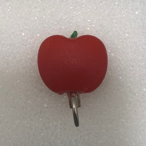 May include: A red apple-shaped hook with a silver metal loop. The hook is made of plastic and has a green stem.