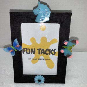 May include: A black picture frame with a white background and the text "FUN TACKS BY: HOME DISTINCTIVES". The frame is decorated with blue and pink butterfly and flower shapes and a blue unicorn head.