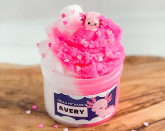 Könnte beinhalten: Ein rosa und weißer Schleim mit einem rosa Axolotl-Anhänger in einem durchsichtigen Glas mit einem Etikett, auf dem "Hello my name is Avery" steht. Der Schleim ist mit rosa Herzstreuseln bedeckt.