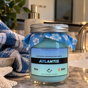 May include: A 9oz soy blend candle in a clear glass jar with a silver metal lid. The candle wax is light blue, and the jar features a blue and white floral fabric topper. The label reads Atlantis, Driftwood, Cedarwood and Amber, 9oz, 60H.
