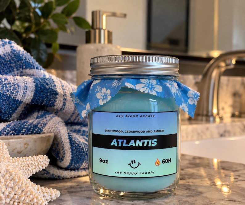 May include: A 9oz soy blend candle in a clear glass jar with a silver metal lid. The candle wax is light blue, and the jar features a blue and white floral fabric topper. The label reads Atlantis, Driftwood, Cedarwood and Amber, 9oz, 60H.