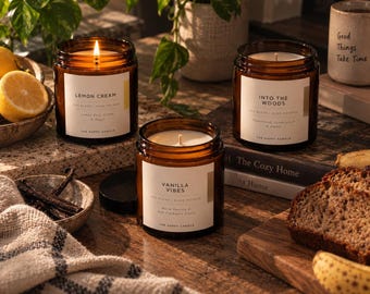 May include: Three amber glass jar candles with white labels. One candle is lit. Labels read Lemon Cream, Into The Woods, and Vanilla Vibes. The candles are soy blend and hand poured by The Happy Candle. The jars have black lids.
