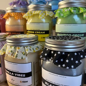 May include: A collection of glass jar candles with metal lids topped with patterned fabric. Labels feature text including 'Vanilla Vibes', 'Black Tide', 'Lemon Cream', and 'Bamboo Grove', with '60H' and '9oz' markings visible on the front labels.