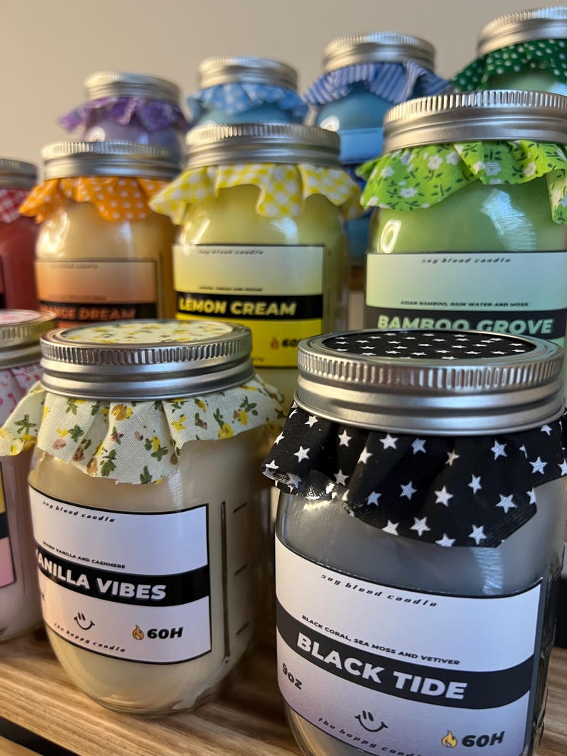 May include: A collection of glass jar candles with metal lids topped with patterned fabric. Labels feature text including 'Vanilla Vibes', 'Black Tide', 'Lemon Cream', and 'Bamboo Grove', with '60H' and '9oz' markings visible on the front labels.