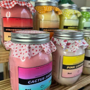 May include: A collection of glass jar candles with silver lids, each topped with a patterned fabric cover. The front labels display scent names like Cactus Jade, Pink Lemonade, and Strawberry Cheesecake, with a 9oz weight clearly printed on each.