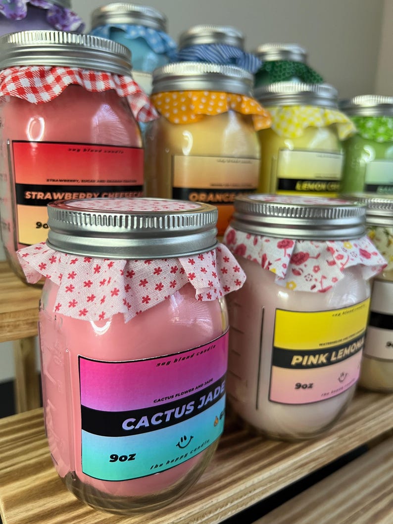 May include: A collection of glass jar candles with silver lids, each topped with a patterned fabric cover. The front labels display scent names like Cactus Jade, Pink Lemonade, and Strawberry Cheesecake, with a 9oz weight clearly printed on each.
