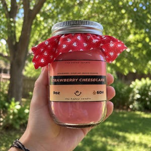 May include: A 9oz soy blend candle in a clear glass jar with a silver lid and a red fabric topper featuring a white heart pattern. The label reads STRAWBERRY CHEESECAKE, STRAWBERRY, SUGAR AND GRAHAM CRACKER, 9oz, 60H, and the happy candle.
