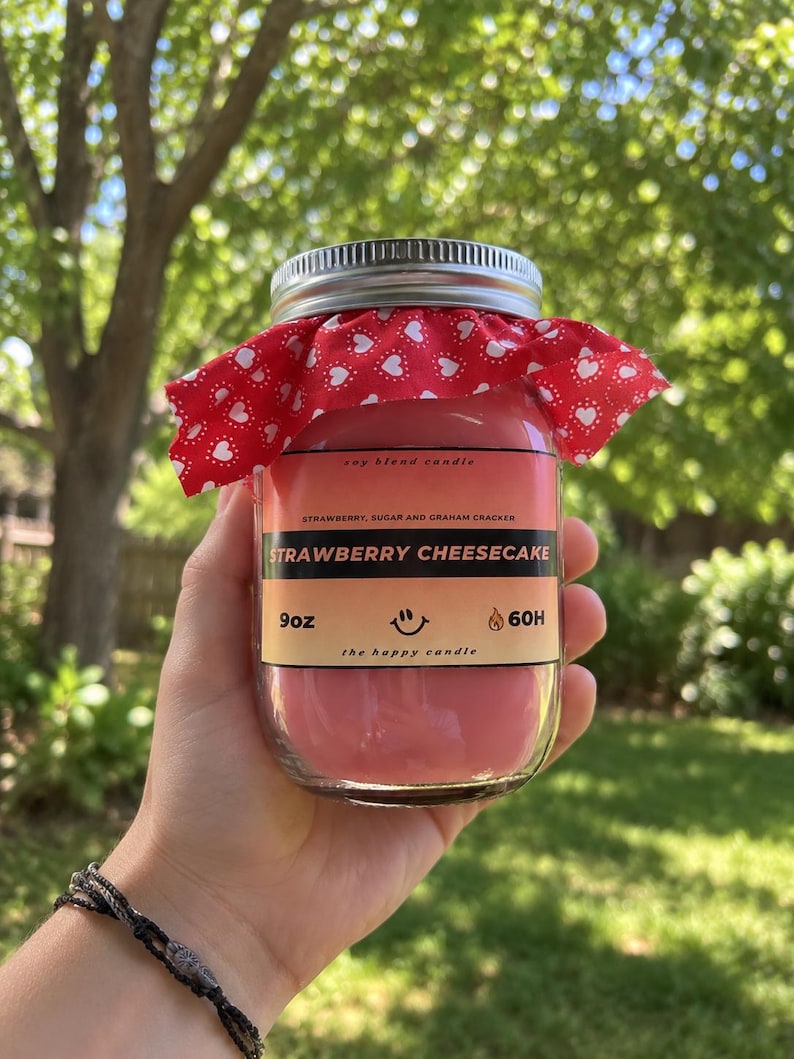 May include: A 9oz soy blend candle in a clear glass jar with a silver lid and a red fabric topper featuring a white heart pattern. The label reads STRAWBERRY CHEESECAKE, STRAWBERRY, SUGAR AND GRAHAM CRACKER, 9oz, 60H, and the happy candle.