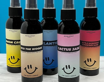 May include: Five black spray bottles with colorful labels. The labels read "Lemon Cream", "Into the Woods", "Atlantis", "Cactus Jade", and "Strawberry Cheesecake". Each label features a smiling face and the text "The Happy Candle".