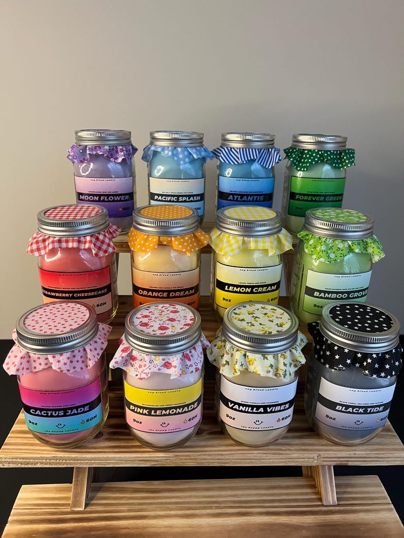 May include: A collection of eleven glass jar candles with metal lids, each topped with a patterned fabric cover. The labels display names like Moon Flower, Pacific Splash, Atlantis, Forever Green, Strawberry Cheesecake, Orange Dream, Lemon Cream, Bamboo Grove, Cactus Jade, Pink Lemonade, Vanilla Vibes, and Black Tide, with each jar marked as 9oz.
