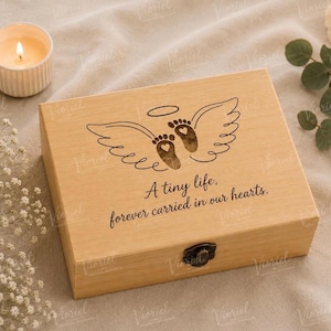 May include: A rectangular light-colored wooden keepsake box with a metal latch. The lid features a laser-engraved design of angel wings with baby footprints containing small hearts, and the text: A tiny life, forever carried in our hearts.