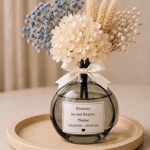 May include: A round, gray glass vase holding a floral arrangement of blue, cream, and beige dried flowers. The vase has a white label with the text "Forever in our hearts." and a name and dates. A white ribbon is tied around the neck.