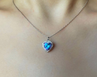 May include: A silver chain necklace with a heart-shaped pendant. The pendant features a large blue gemstone surrounded by smaller clear gemstones.