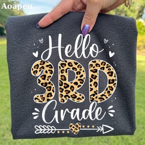 May include: A dark charcoal gray t-shirt featuring the white text Hello and Grade with a leopard print pattern filling the large 3RD characters. The design includes small white heart icons and a horizontal arrow graphic with a leopard print heart.