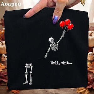 May include: A black t-shirt featuring a white graphic of a skeleton torso holding three red balloons, with the skeleton's legs separated below. The text "Well, shit..." is printed in white below the skeleton torso on the front of the garment.