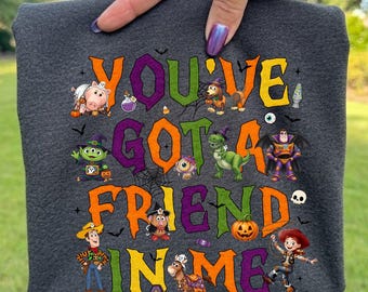 Può includere: Una t-shirt grigio scuro con il testo YOU'VE GOT A FRIEND IN ME in lettere arancioni, viola e verdi. Il design include personaggi dei cartoni animati in costumi di Halloween, ragnatele, pipistrelli, una zucca e un'immagine di un teschio.