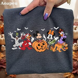 Puede incluir: Una camiseta gris oscuro con un estampado gráfico de personajes de Disney con disfraces de Halloween, incluyendo a Goofy como momia, el Pato Donald como diablo, Mickey Mouse en una calabaza, Minnie Mouse como esqueleto, Daisy como bruja y Pluto.
