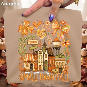 Pode incluir: Um artigo de tecido bege com um gráfico colorido de uma pequena cidade com edifícios, uma torre de água e uma placa com o número 19008. O design inclui flores laranja e amarelas, um laço grande e o texto SMALL TOWN FALL em letras laranja.
