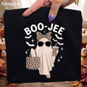 May include: A black t-shirt featuring a white graphic of a ghost wearing leopard print sunglasses and a matching bow. The ghost holds a leopard print tote bag and a matching cup. The text BOO-JEE is printed above the ghost with small white bats.