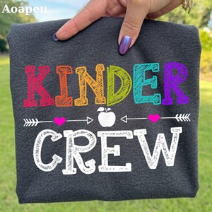 May include: A dark gray t-shirt featuring the text "KINDER" in multicolored, sketch-style letters, followed by a white apple icon flanked by two small pink hearts and horizontal arrows, and the word "CREW" in large, white, sketch-style letters.