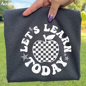 Puede incluir: Una camiseta gris oscuro con un gráfico blanco de una manzana con patrón de cuadros. El texto "LET'S LEARN TODAY" está impreso en disposición circular alrededor de la manzana, junto a pequeños iconos de estrellas a cuadros.