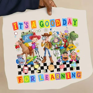 May include: A light beige fabric featuring a colourful graphic of Toy Story characters including Woody, Buzz Lightyear, Jessie, Rex, and Mr. Potato Head with school supplies. The text reads IT'S A GOOD DAY FOR LEARNING in multi-coloured block letters.