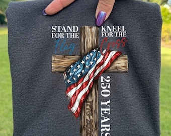 May include: A dark charcoal gray t-shirt featuring a graphic of a wooden cross draped with an American flag. The text reads STAND FOR THE FLAG KNEEL FOR THE CROSS 250 YEARS 1776-2026. The design is centered on the fabric with blue and red lettering.