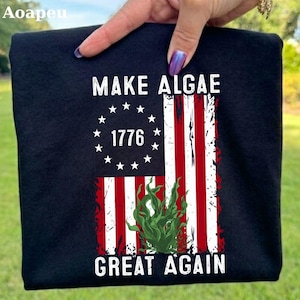 Könnte beinhalten: Ein schwarzes T-Shirt mit einem Grafikdruck einer stilisierten amerikanischen Flagge mit roten und weißen Streifen und einem Kreis aus weißen Sternen um die Zahl 1776. Der Text "MAKE ALGAE" steht darüber und "GREAT AGAIN" darunter.
