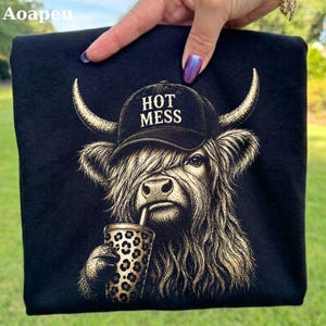 May include: A black t-shirt featuring a graphic of a Highland cow wearing a black baseball cap with the text HOT MESS printed in white. The cow has long shaggy fur and is holding a cup with a leopard print pattern in its hoof. The design is gold.