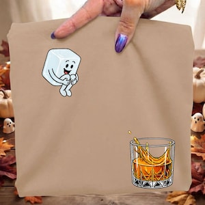 Puede incluir: Una camiseta de color tostado con un gráfico de dibujos animados de un cubito de hielo sonriente con brazos y piernas en la parte superior izquierda y un vaso de líquido ámbar con efecto de salpicadura en la parte inferior derecha.