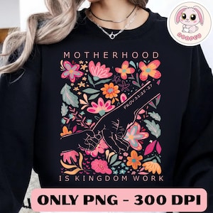May include: Black sweatshirt with a floral design and the words "Motherhood Is Kingdom Work." The design includes colorful flowers and two hands holding each other. The text "Prov. 31:25-27" is also included.