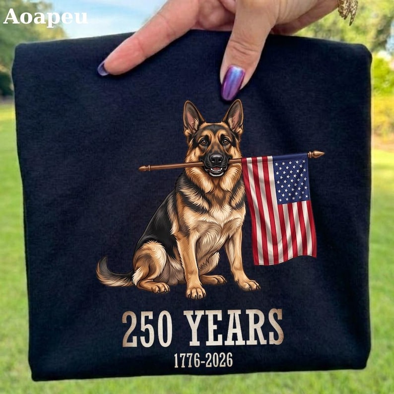 May include: A black t-shirt featuring a graphic of a German Shepherd dog holding a United States flag in its mouth. Below the dog, the text reads 250 YEARS and 1776-2026 in a bold, light-colored font. The design is centered on the fabric.