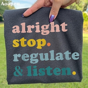 Könnte beinhalten: Ein dunkel anthrazitfarbenes T-Shirt mit dem Text "alright stop. regulate & listen." in gestapeltem Layout. Das Wort "alright" ist hellrosa, "stop." ist gelb, "regulate" ist hellblau und "& listen." ist in der Farbe Petrol.
