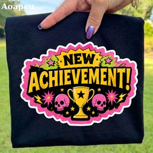 May include: A black t-shirt featuring a graphic design with the text NEW ACHIEVEMENT! in yellow letters. The design includes a central yellow trophy, two pink skulls on either side, and decorative pink and yellow starbursts on a pink background.