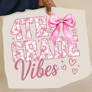May include: A light beige t-shirt featuring the text 4TH GRADE VIBES in a white block font with a pink floral pattern. A large, glossy pink bow is positioned above the word GRADE, accompanied by small pink hearts and four-pointed star accents.