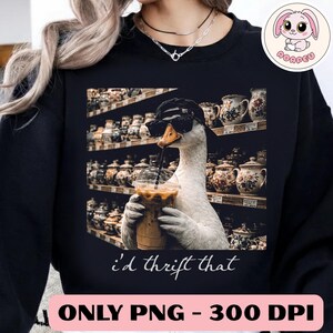 Pode incluir: Camisola preta com um desenho de um ganso usando óculos de sol e um chapéu, bebendo café gelado. O ganso está em frente a prateleiras de cerâmica vintage. O texto "i'd thrift that" também está presente.