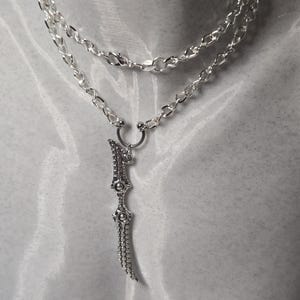 May include: A silver chain necklace with a silver sword pendant. The sword is detailed with small circles and has a pointed tip.