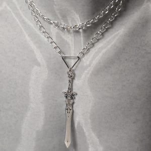 May include: A silver chain necklace with a silver sword pendant. The sword has intricate details and a pointed tip.