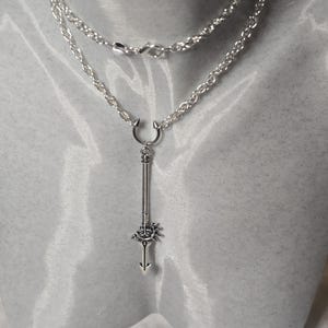 May include: A silver chain necklace with a silver pendant featuring a long, pointed arrow with a circular design at the top. The arrow is attached to a horseshoe-shaped piece at the top of the chain.