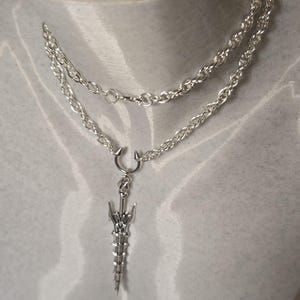 May include: A silver chain necklace with a silver sword pendant. The sword has a detailed design and a pointed tip.