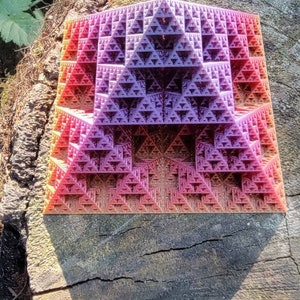 May include: A geometric, three-dimensional sculpture with a fractal design. The sculpture is composed of numerous small triangles, with colors ranging from red to purple. The object is resting on a tree stump.
