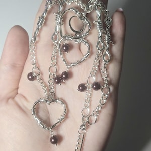 Op de afbeelding: Een zilveren ketting met meerdere hartvormige bedels van prikkeldraad. De ketting heeft ook bruine parel accenten.