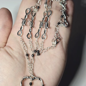 Op de afbeelding: Een zilveren ketting met een hartvormige hanger gemaakt van prikkeldraad. De ketting heeft ook meerdere strengen zilveren ketting en zwarte kralen.